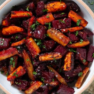 Caramelized roasted beets and carrots with balsamic vinaigrette garnished with chopped fresh parsley and flaky sea salt in a white bowl.