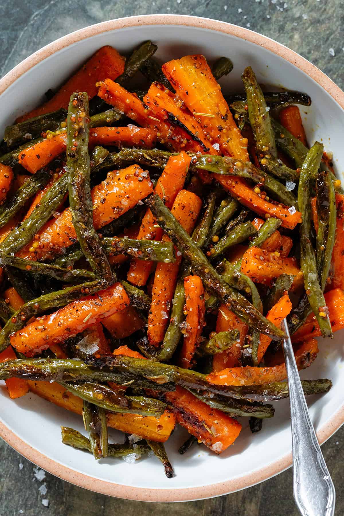 Roasted carrots and green beans with whole mustard vinaigrette in a white bowl with a beige rim with a fork inserted on the right.