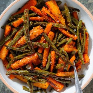 Roasted carrots and green beans with whole mustard vinaigrette in a white bowl with a beige rim with a fork inserted on the right.