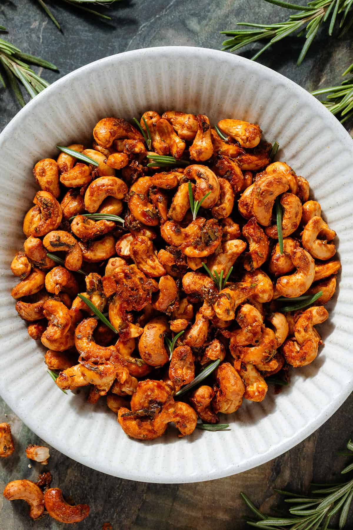 Golden roasted cashews with rosemary in a white ceramic bowl garnished with fresh rosemary.