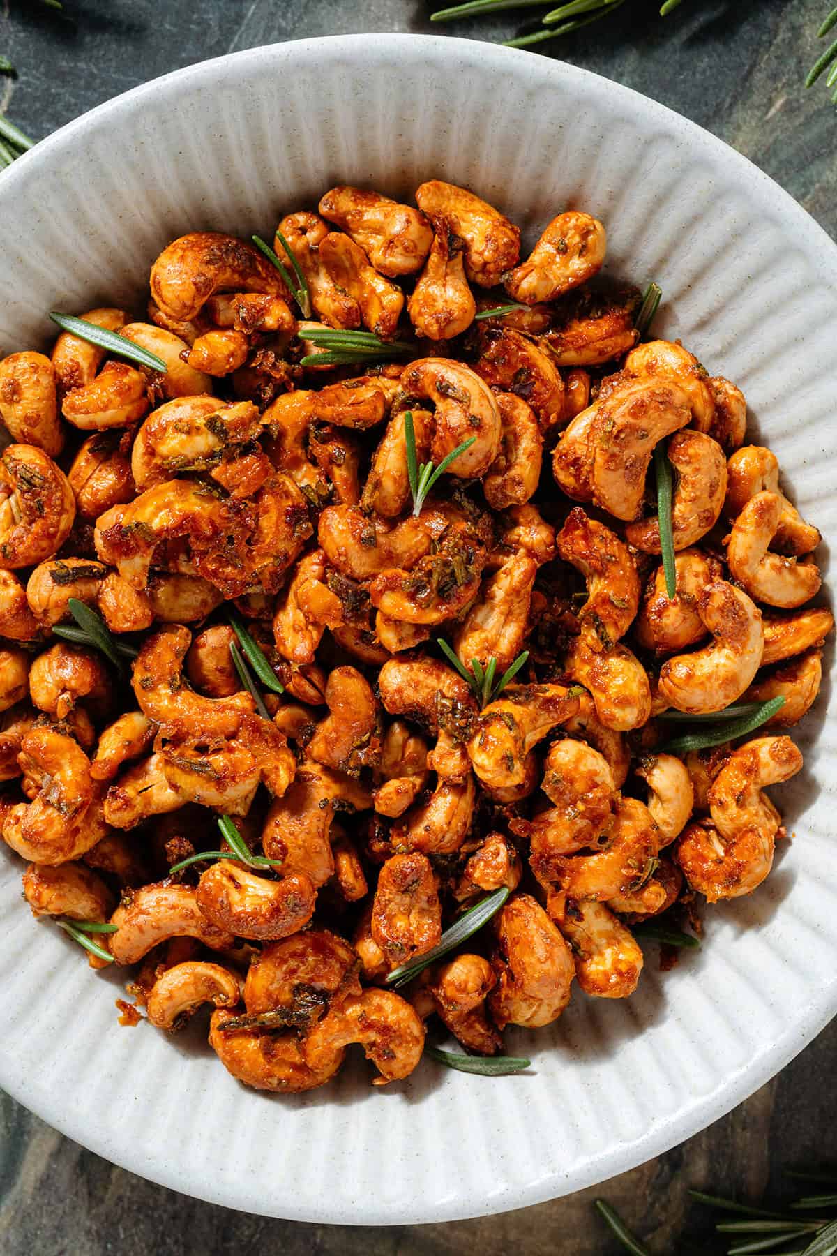 Golden roasted cashews with rosemary in a white ceramic bowl garnished with fresh rosemary.