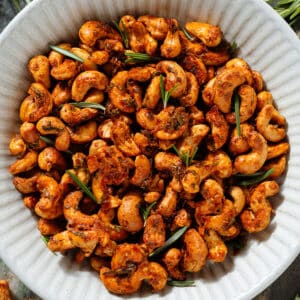 Golden roasted cashews with rosemary in a white ceramic bowl garnished with fresh rosemary.
