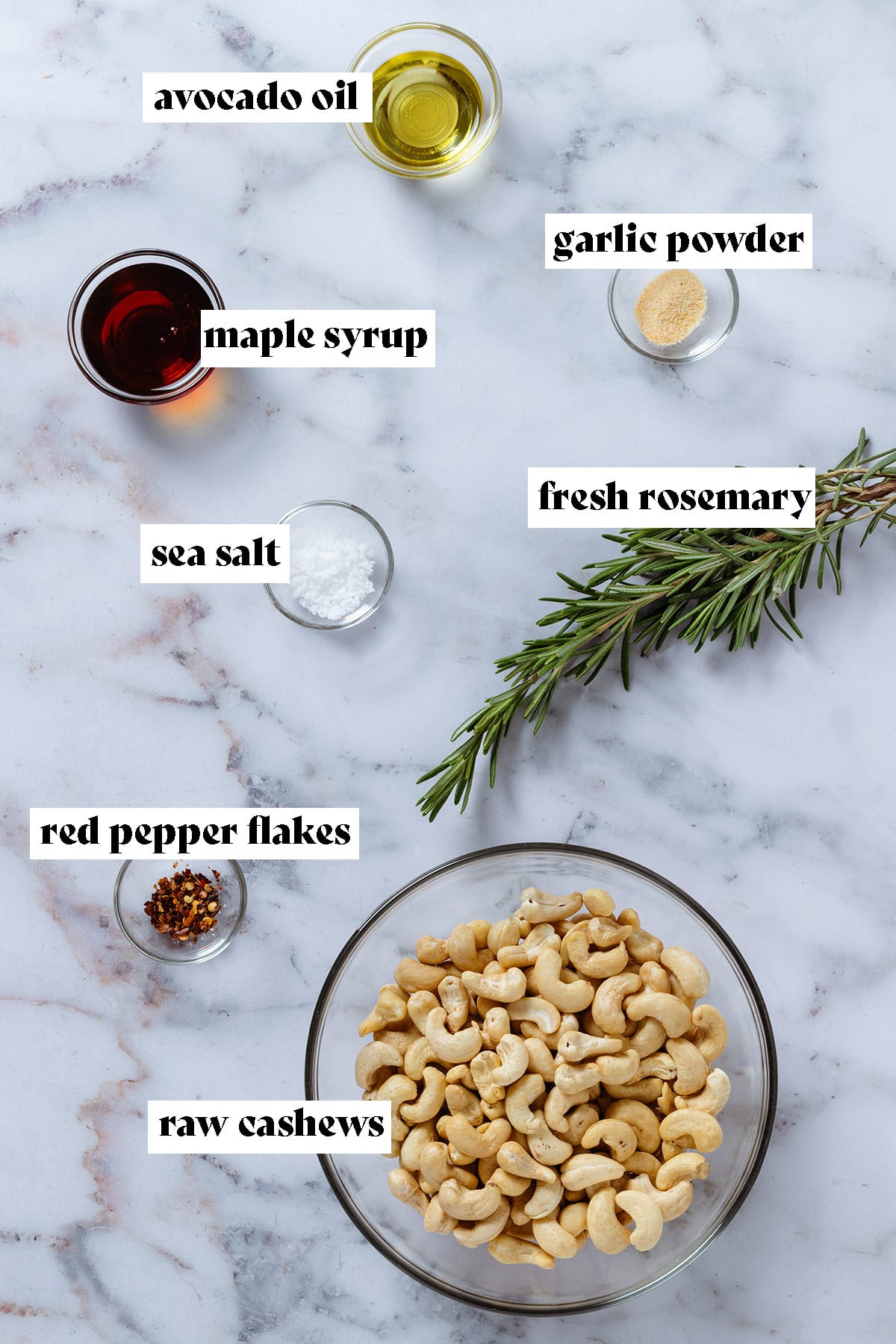 Ingredients like cashews, maple syrup, avocado oil, fresh rosemary, and spices all laid out in small glass bowls on a marble background with text overlay.