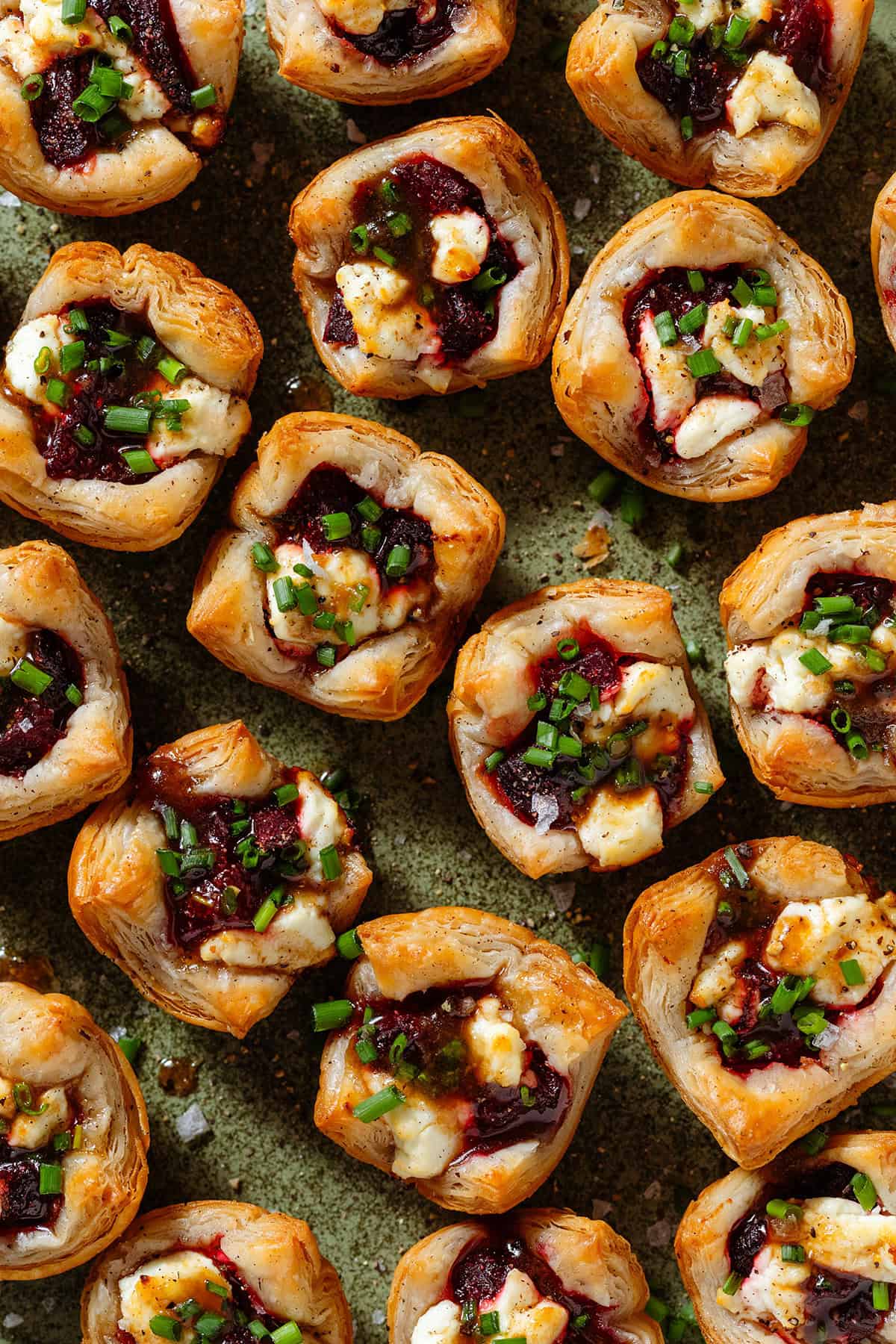 Lots of puff pastry cups with diced beets and slightly melted goat cheese topped with balsamic vinaigrette, black pepper, and chopped chives on a green plate.
