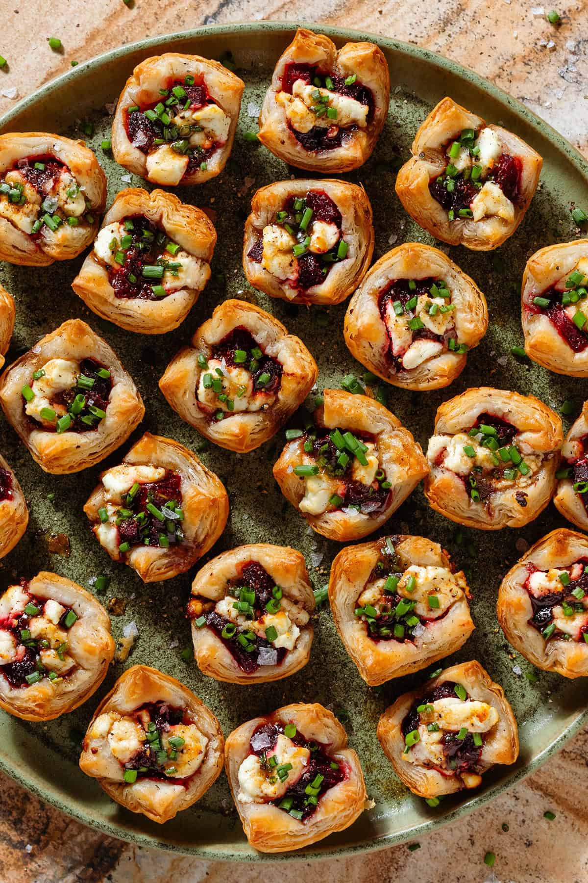 Lots of puff pastry cups with diced beets and slightly melted goat cheese topped with balsamic vinaigrette and chopped chives on a green plate.