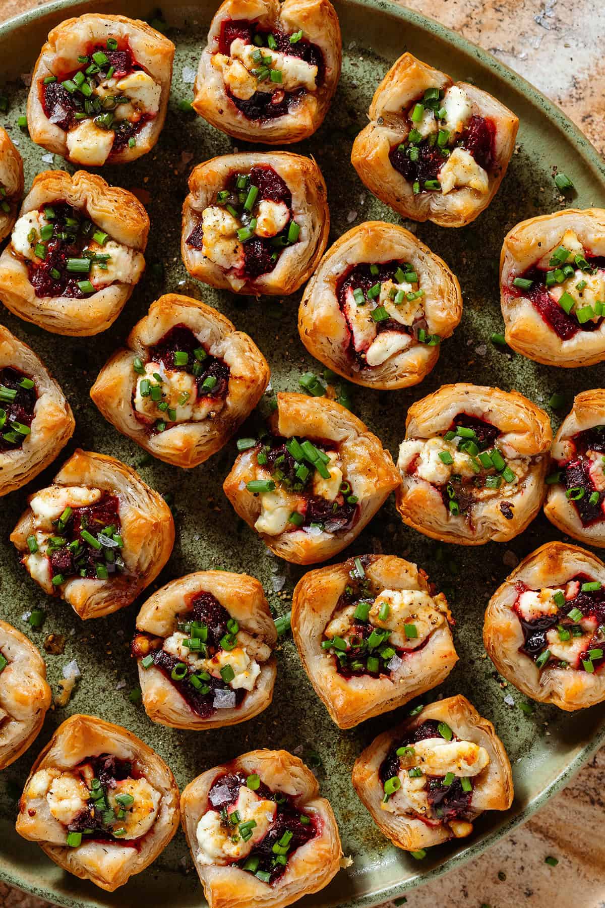 Lots of puff pastry cups with diced beets and slightly melted goat cheese topped with balsamic vinaigrette and chopped chives on a green plate.