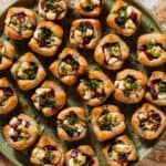 Lots of puff pastry cups with diced beets and slightly melted goat cheese topped with balsamic vinaigrette and chopped chives on a green plate.