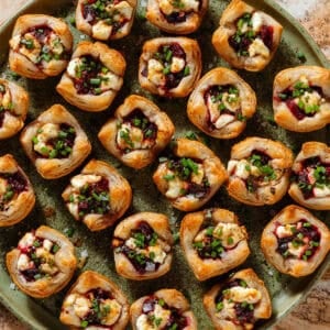 Lots of puff pastry cups with diced beets and slightly melted goat cheese topped with balsamic vinaigrette and chopped chives on a green plate.