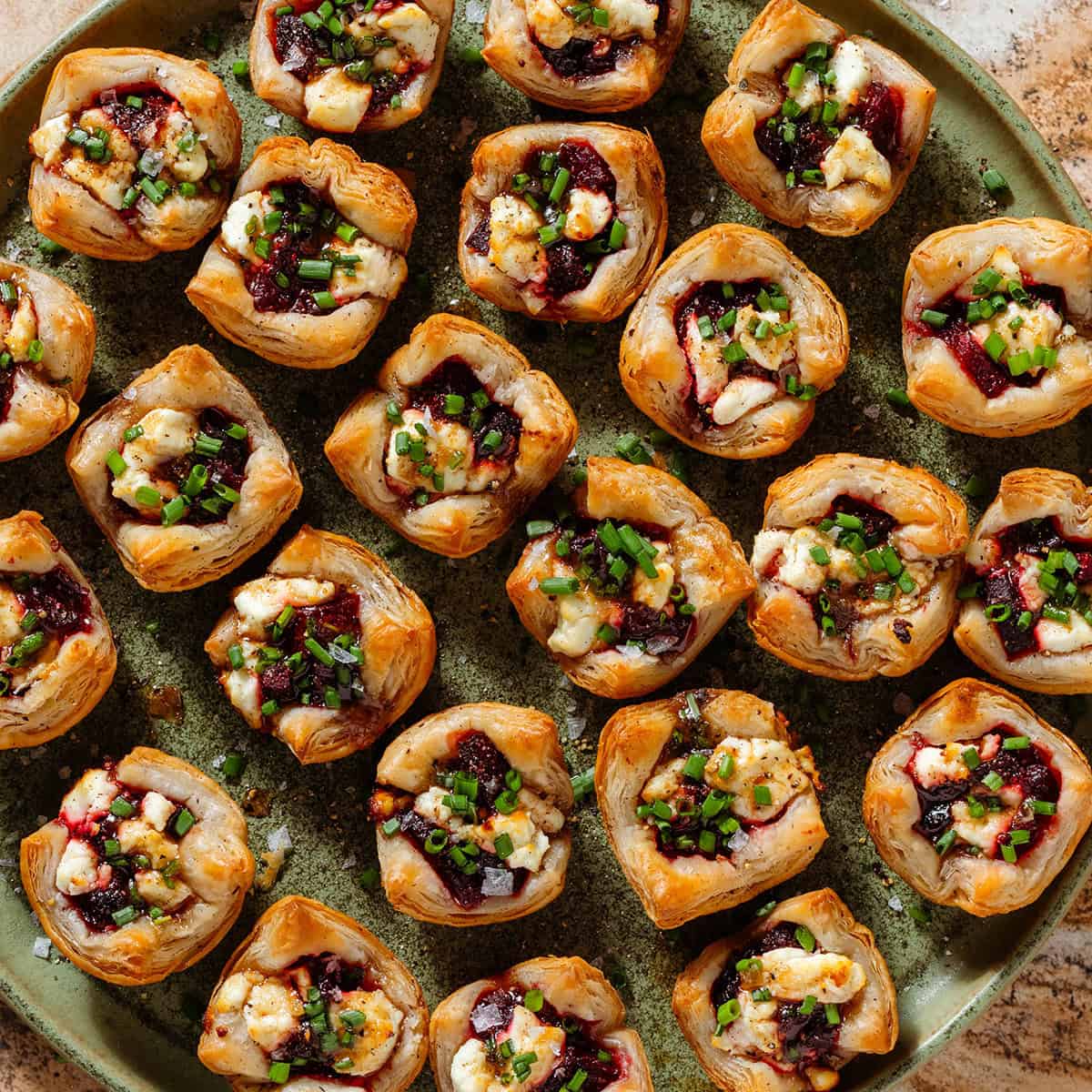 Lots of puff pastry cups with diced beets and slightly melted goat cheese topped with balsamic vinaigrette and chopped chives on a green plate.
