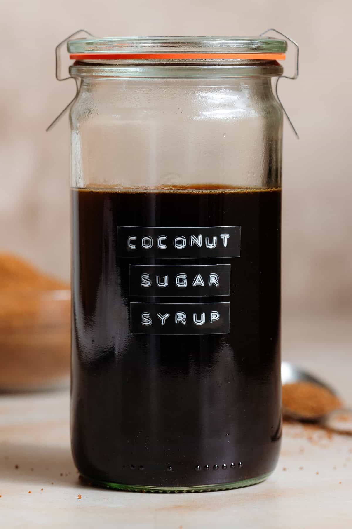 Coconut sugar syrup in a tall glass jar with an embossed label on a beige background with more coconut sugar in the background in a glass bowl.
