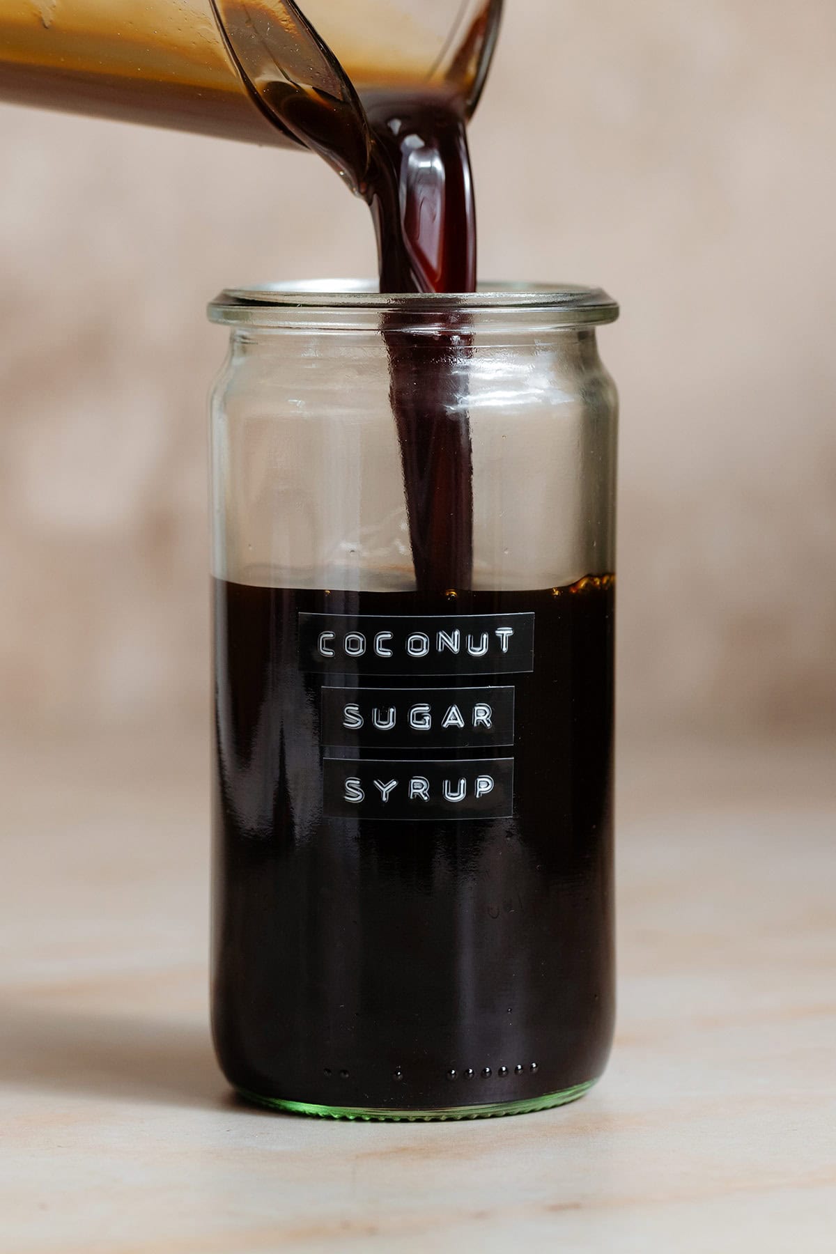 Coconut sugar syrup being poured into a tall glass jar with an embossed label on a beige background.