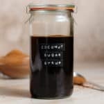 Coconut sugar syrup in a tall glass jar with an embossed label on a beige background with more coconut sugar in the background in a glass bowl.