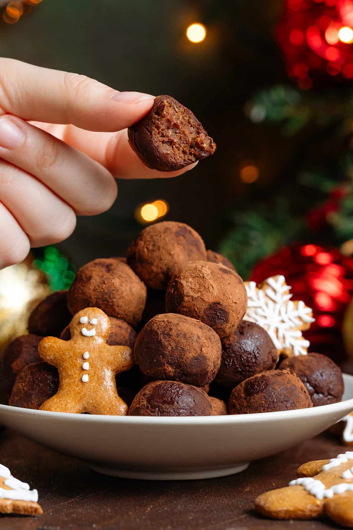 Energy balls rolled in cocoa powder stacked on a small beige plate garnished with mini gingerbread cookies with a hand holding one with a bite taken out of it above them.