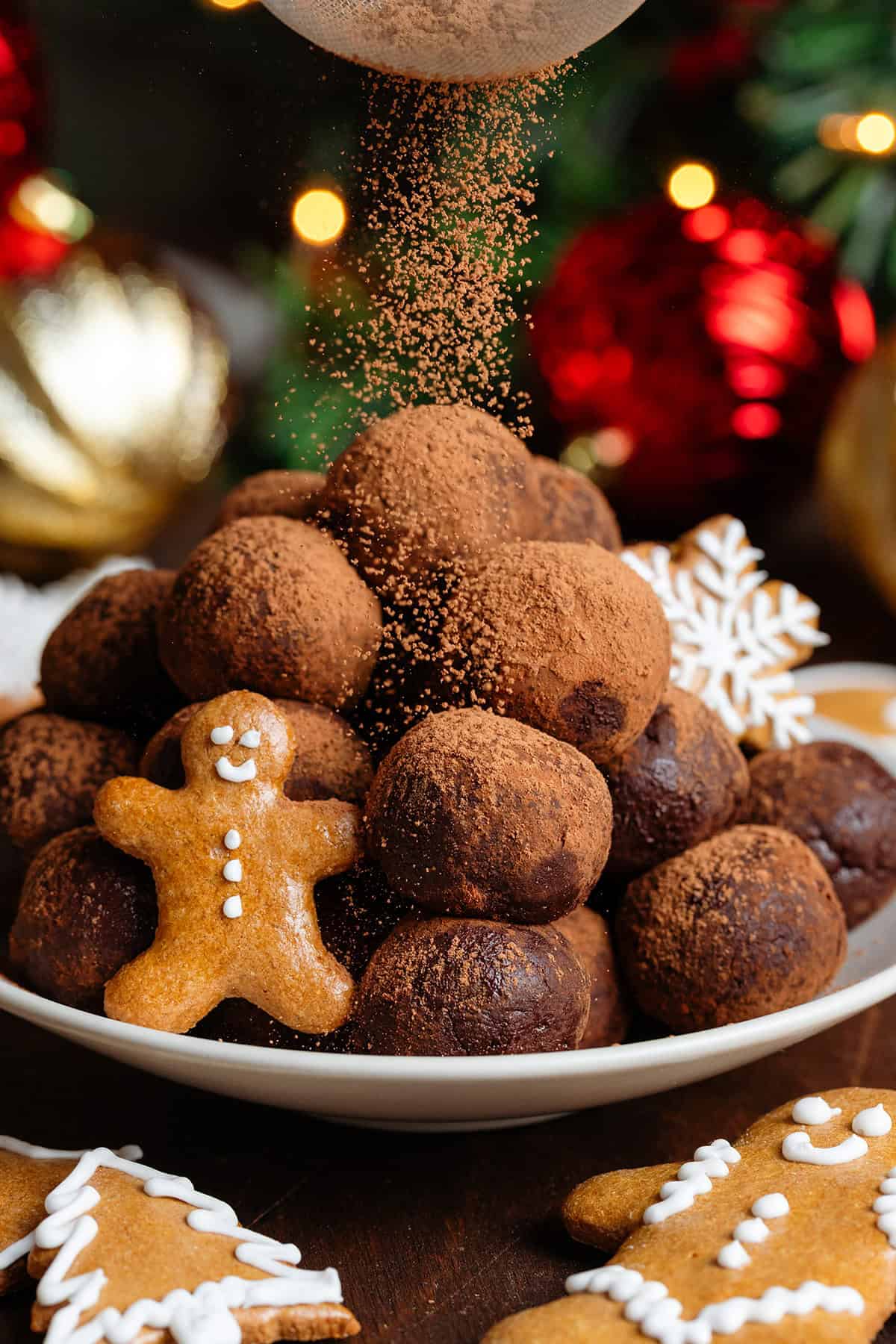Energy balls rolled in cocoa powder stacked on a small beige plate garnished with mini gingerbread cookies and being dusted with more cocoa.