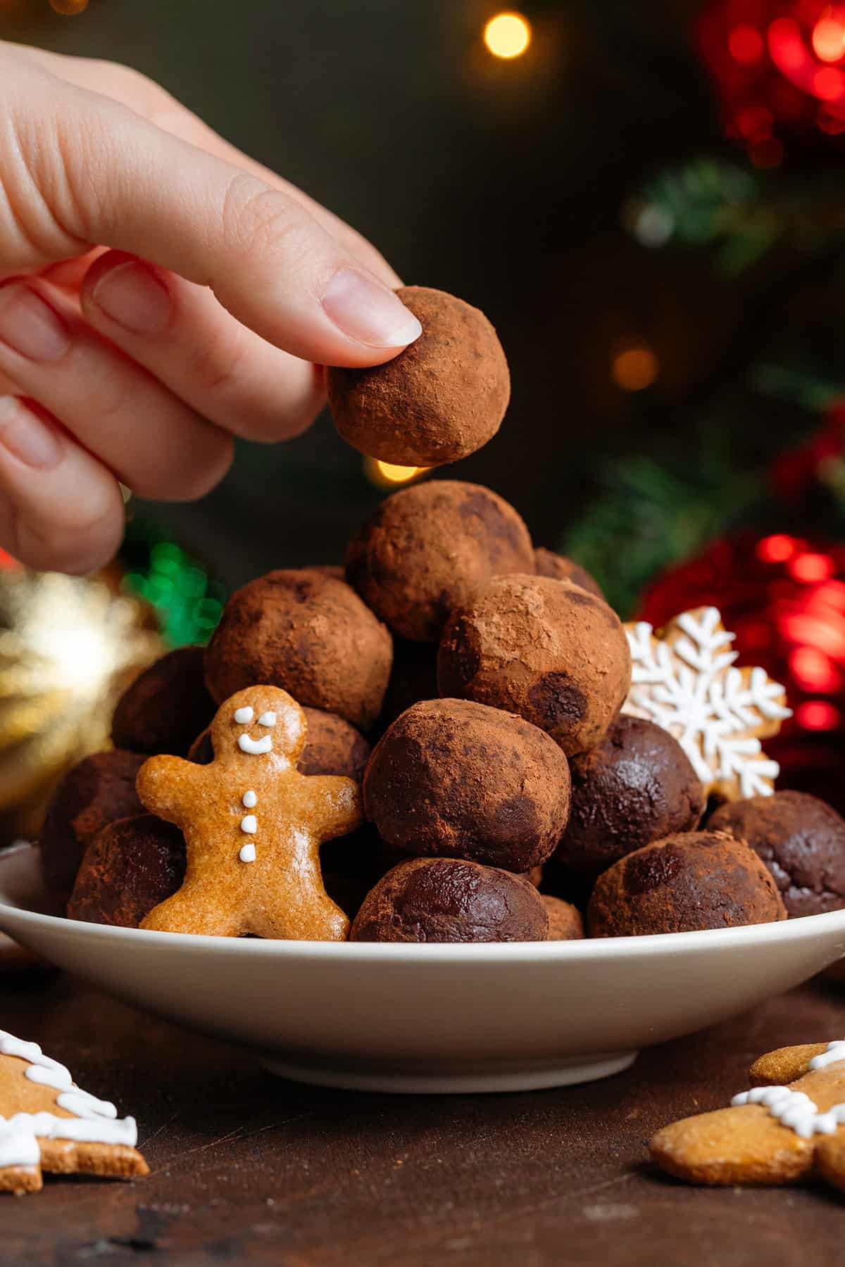Energy balls rolled in cocoa powder stacked on a small beige plate garnished with mini gingerbread cookies with a hand grabbing the one on top.