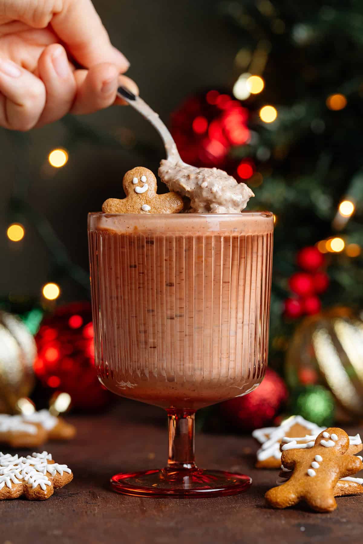 Gingerbread overnight oats in a stemmed red glass garnished with a mini gingerbread man and a hand scooping up the oatmeal with a spoon.