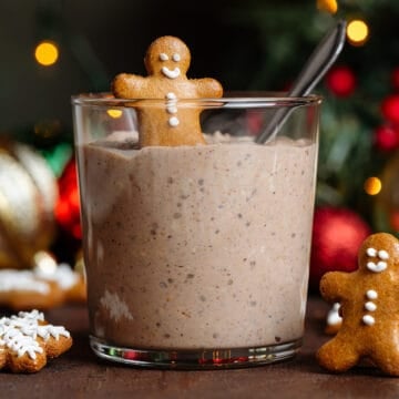 Overnight oats in a short glass topped with a small gingerbread man and a spoon in it with more gingerbread cookies around the glass.