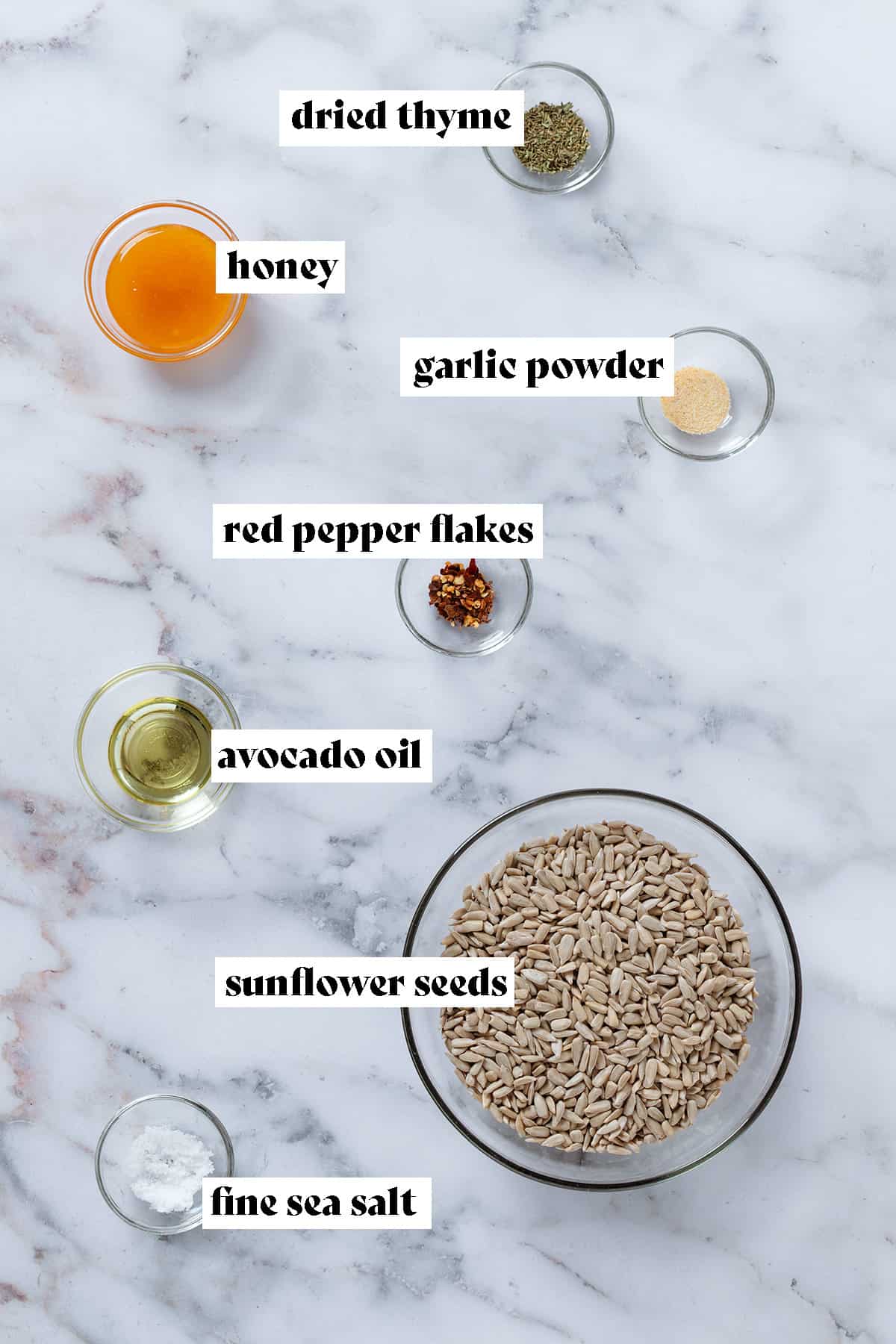 Ingredients like sunflower seeds, avocado oil, honey, and spices in small glass bowls on a marble background with text overlay.