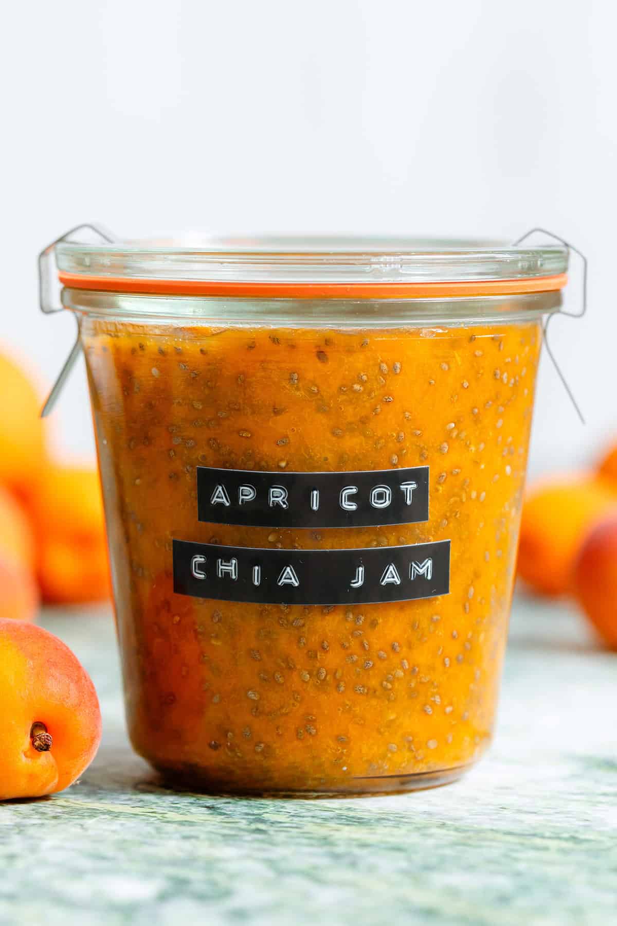 A glass jar with bright orange apricot chia jam with an embossed label and fresh apricots in the background.