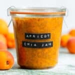 A glass jar with bright orange apricot chia jam with an embossed label and fresh apricots in the background.