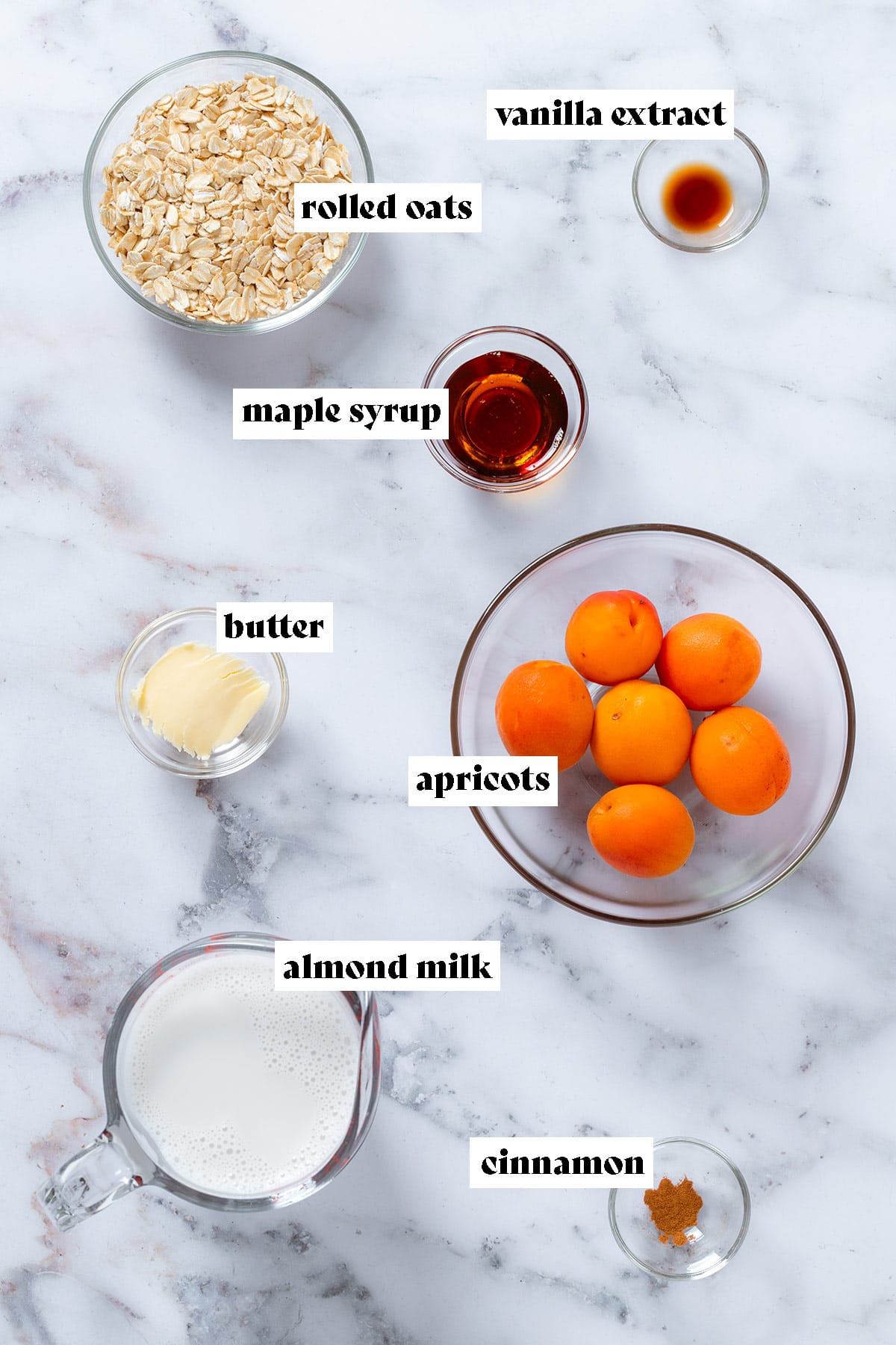 Ingredients like apricots, oats, maple syrup, milk and spices in glass bowls on a marble background with text overlay.