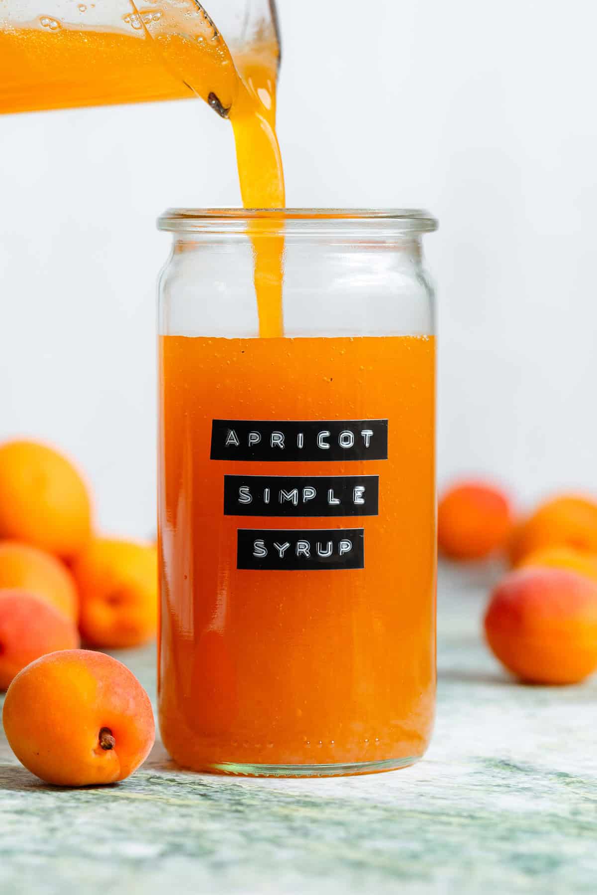 Bright orange apricot syrup being poured into a tall glass jar with an embossed label and apricots in the background.