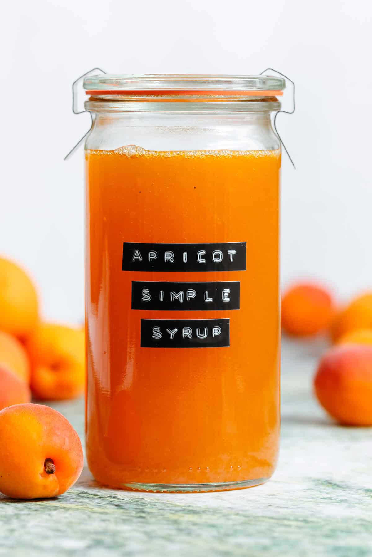 A tall glass jar with bright orange apricot syrup with an embossed label and apricots in the background.