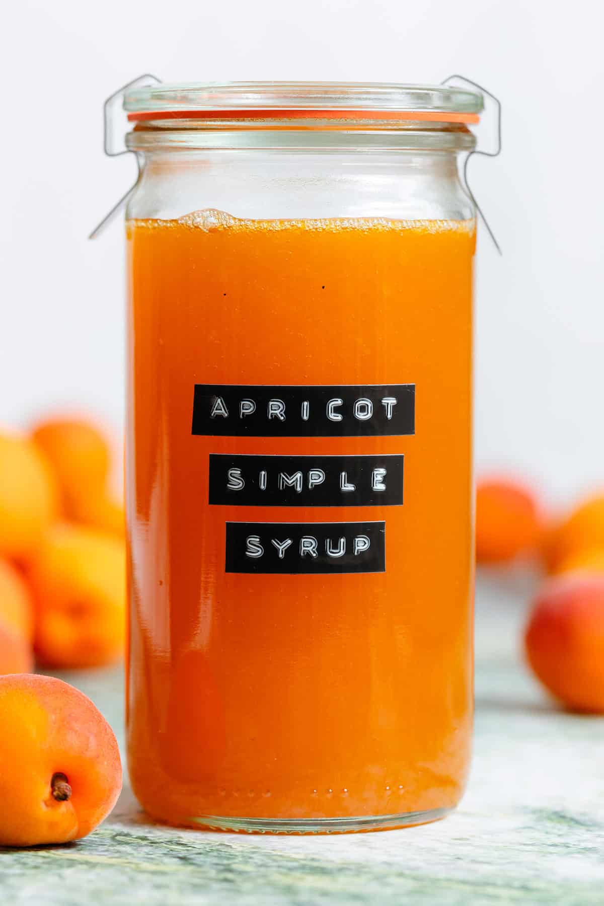 A tall glass jar with bright orange apricot syrup with an embossed label and apricots in the background.