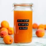 A tall glass jar with bright orange apricot syrup with an embossed label and apricots in the background.