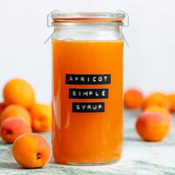 A tall glass jar with bright orange apricot syrup with an embossed label and apricots in the background.