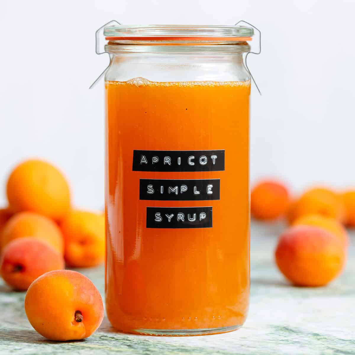 A tall glass jar with bright orange apricot syrup with an embossed label and apricots in the background.