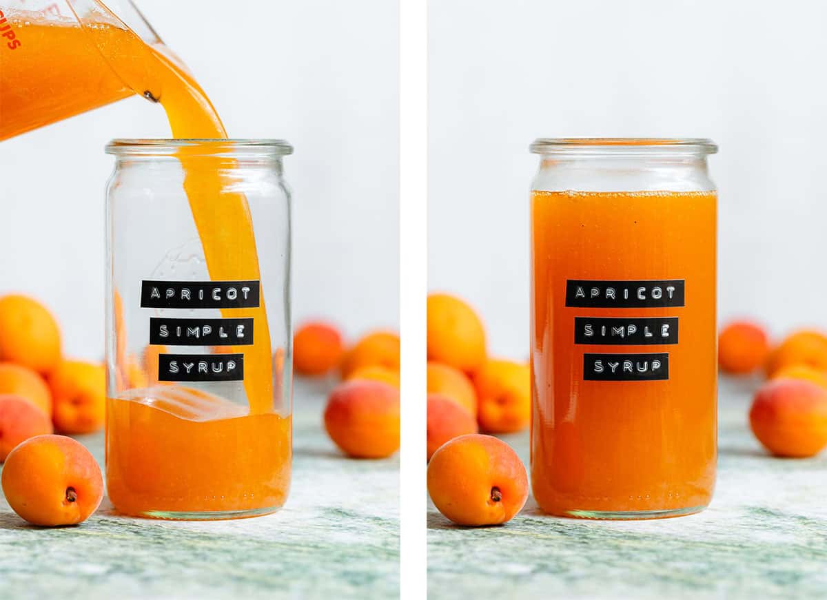 Bright orange apricot syrup being poured into a tall glass jar with an embossed label and apricots in the background.