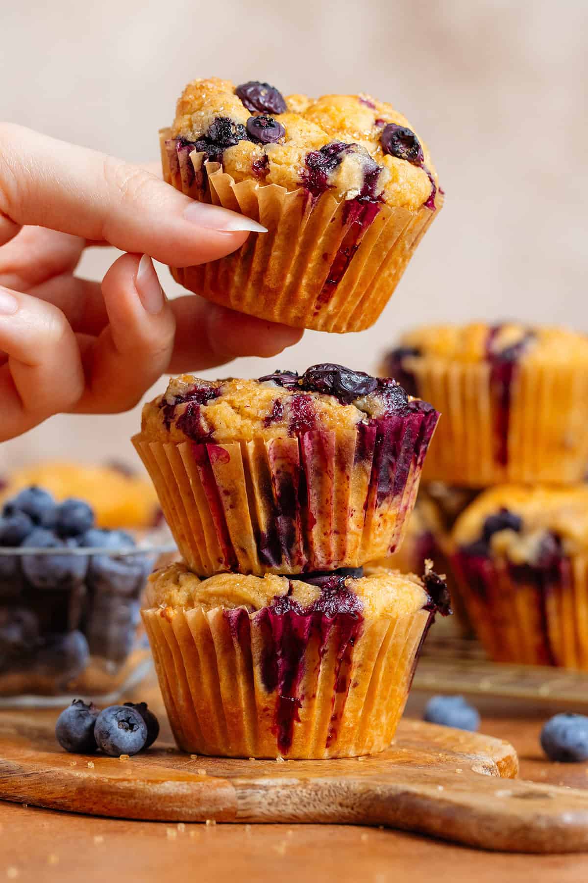 Three blueberry muffins stacked on a small wooden cutting board with a hand grabbing the top one, with fresh blueberries and more muffins in the background.
