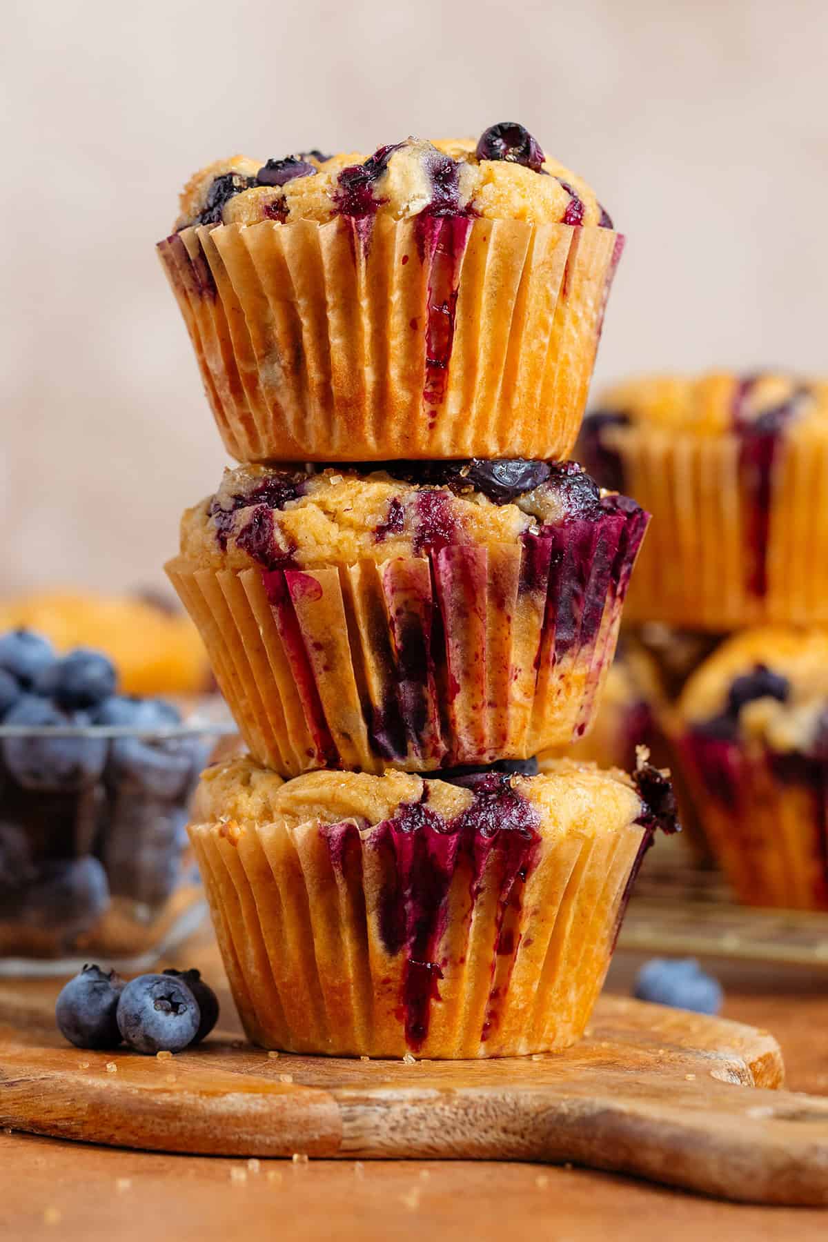 Three blueberry muffins stacked on a small wooden cutting board with fresh blueberries and more muffins in the background.