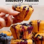 Three blueberry muffins stacked on a small wooden cutting board with fresh blueberries and more muffins in the background.
