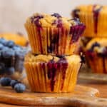 Two blueberry muffins stacked on a small wooden cutting board with fresh blueberries and more muffins in the background.