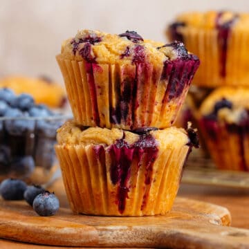 Two blueberry muffins stacked on a small wooden cutting board with fresh blueberries and more muffins in the background.