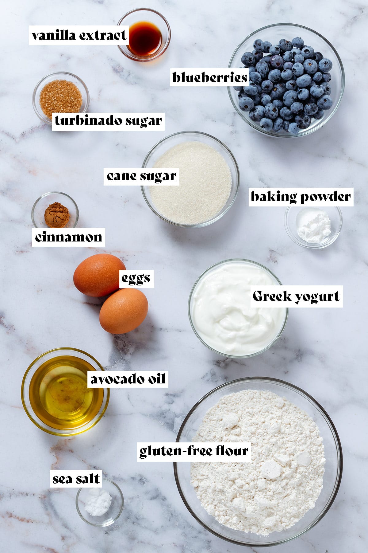 Ingredients like flour, blueberries, sugar, yogurt, oil, and eggs laid out in small glass bowls on marble background with text overlay.