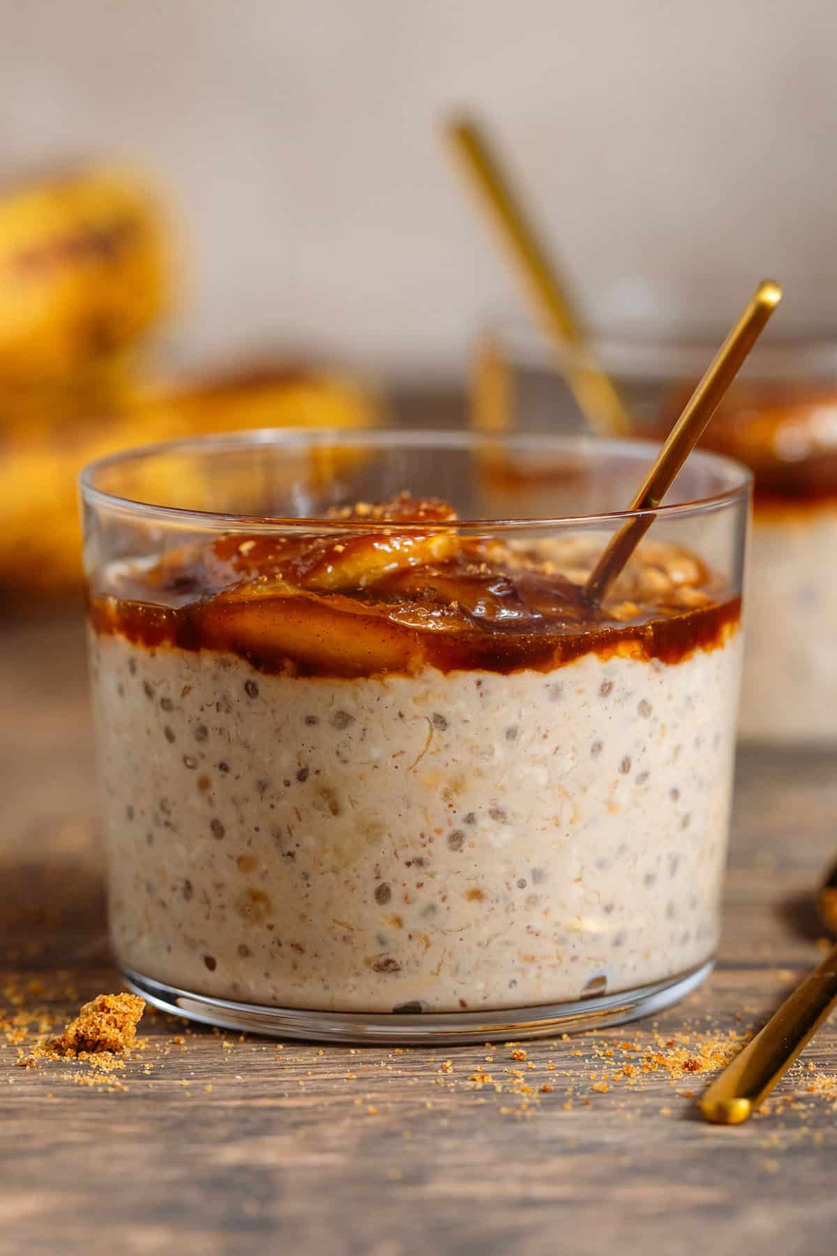 A short glass with brown sugar overnight oats with chia seeds topped with caramelized banana and more sugar with a gold spoon in the oatmeal on the right.