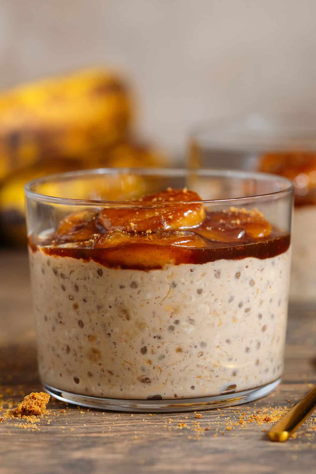 A short glass with brown sugar overnight oats with chia seeds topped with caramelized banana and more sugar with a gold spoon next to the glass.