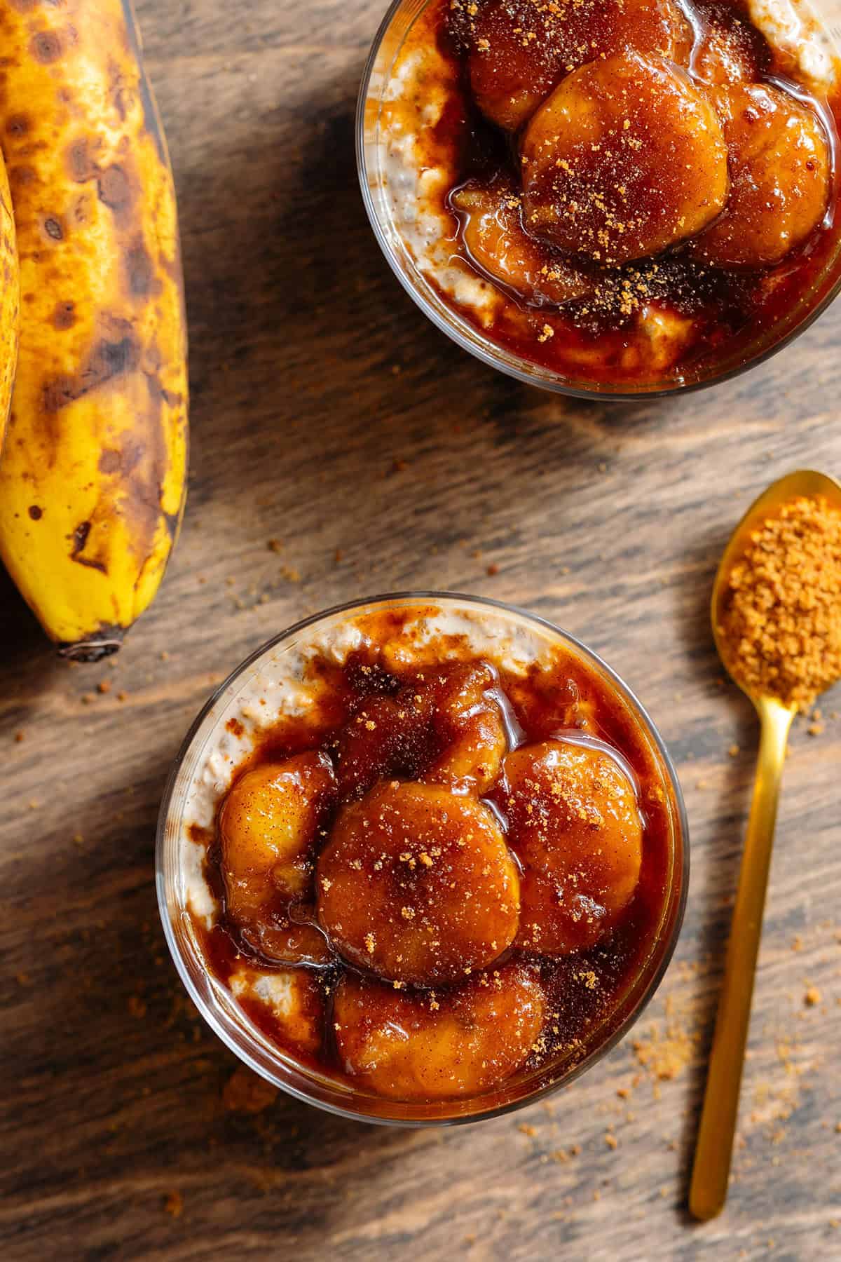 Two short glasses with brown sugar overnight oats topped with caramelized banana and more sugar with a gold spoon next to the one on the bottom.