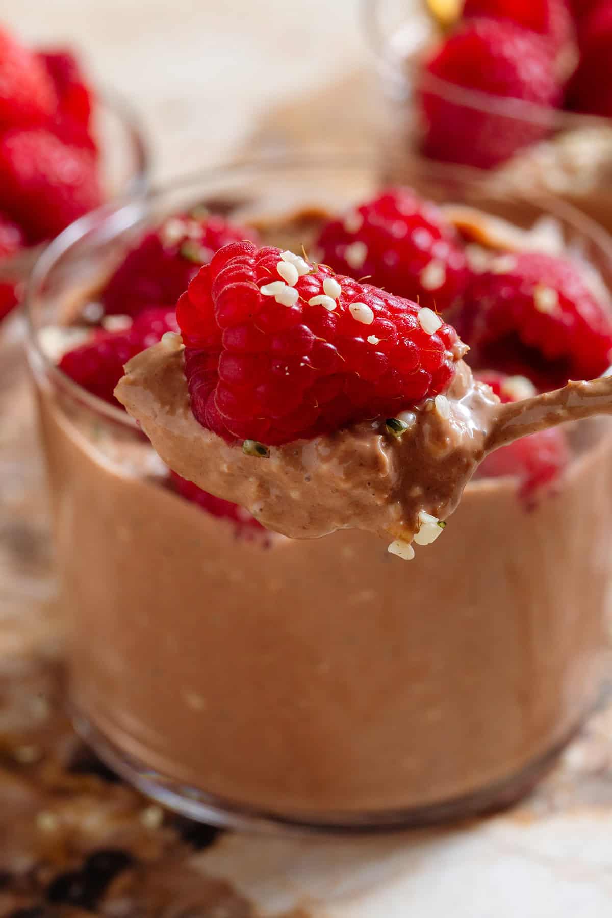 A spoon holding chocolate overnight oats with a raspberry and seeds with more of the oatmeal in a short glass in the background.