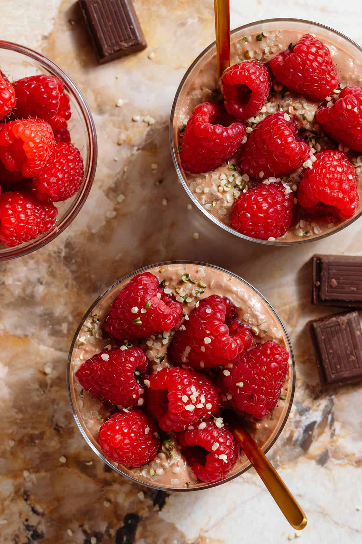 Chocolate overnight oats in two short glasses garnished with fresh raspberries and seeds and with a gold spoons.