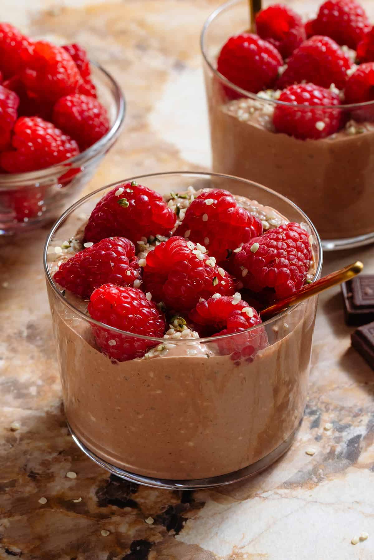 Chocolate overnight oats in two short glasses garnished with fresh raspberries and seeds and with a gold spoons.