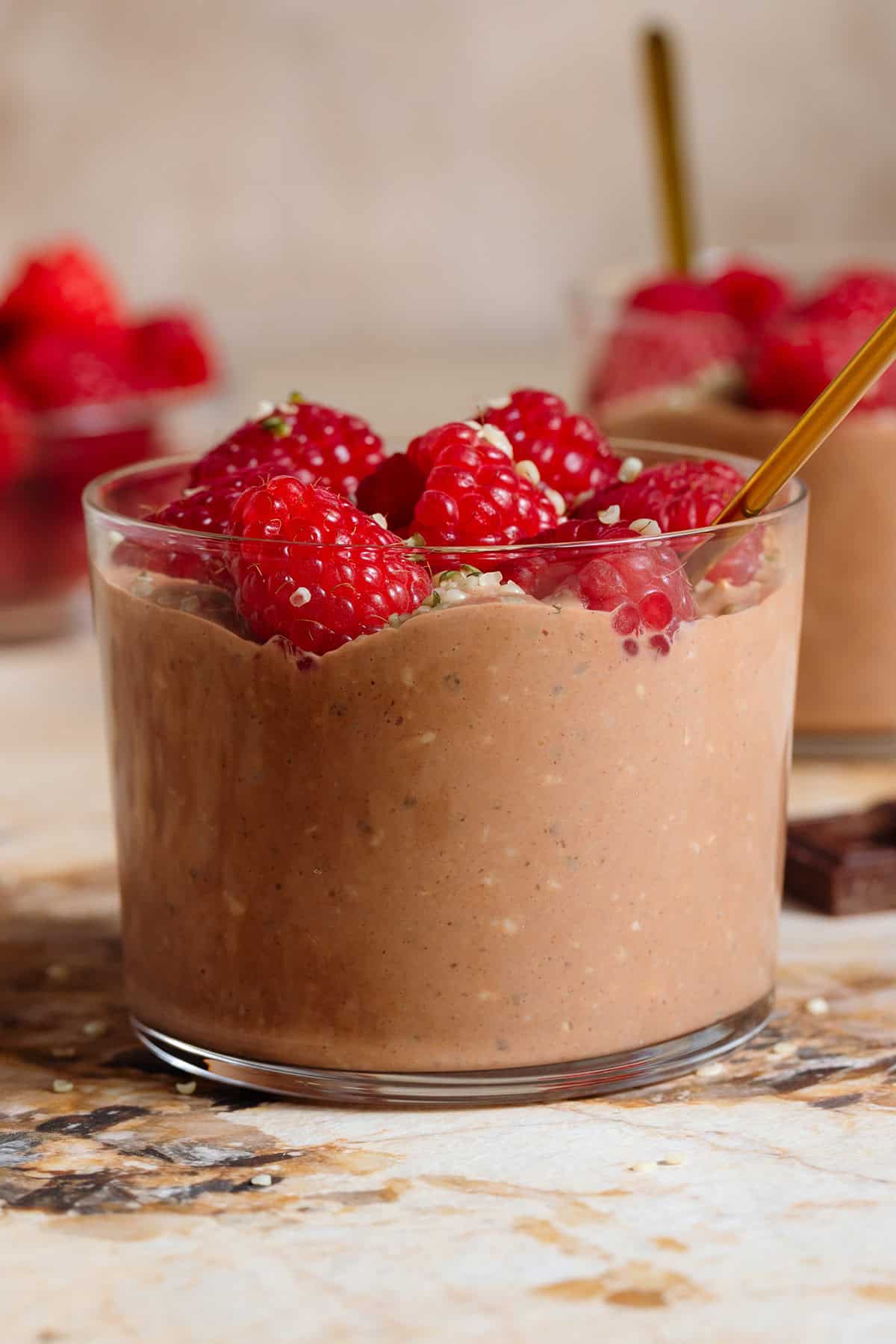 Chocolate overnight oats in a short glasse garnished with fresh raspberries and seeds and with a gold spoon with another glass in the background.