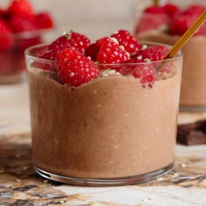 Chocolate overnight oats in a short glass garnished with fresh raspberries and seeds and with a gold spoon.