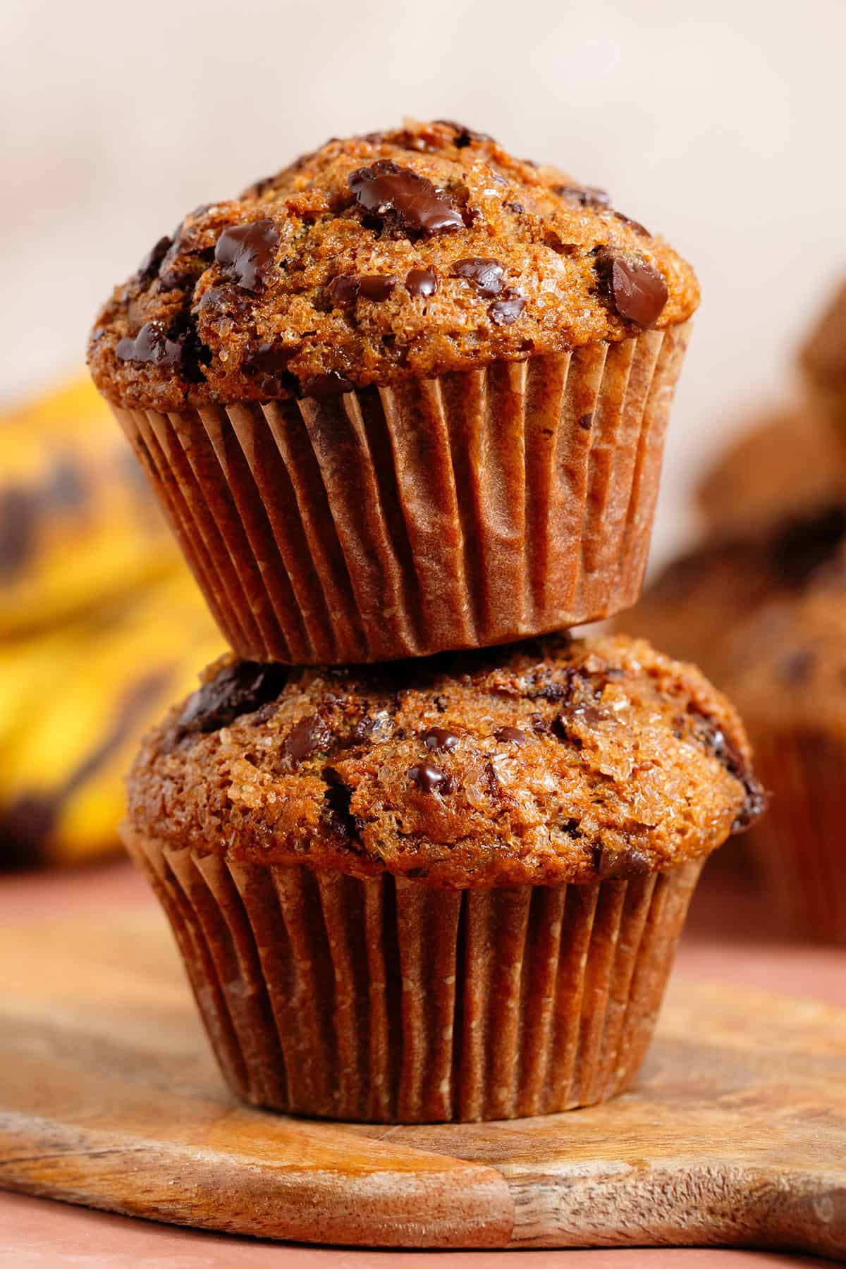 Two chocolate chunk muffins stacked on top of each other on a small wooden cutting board.