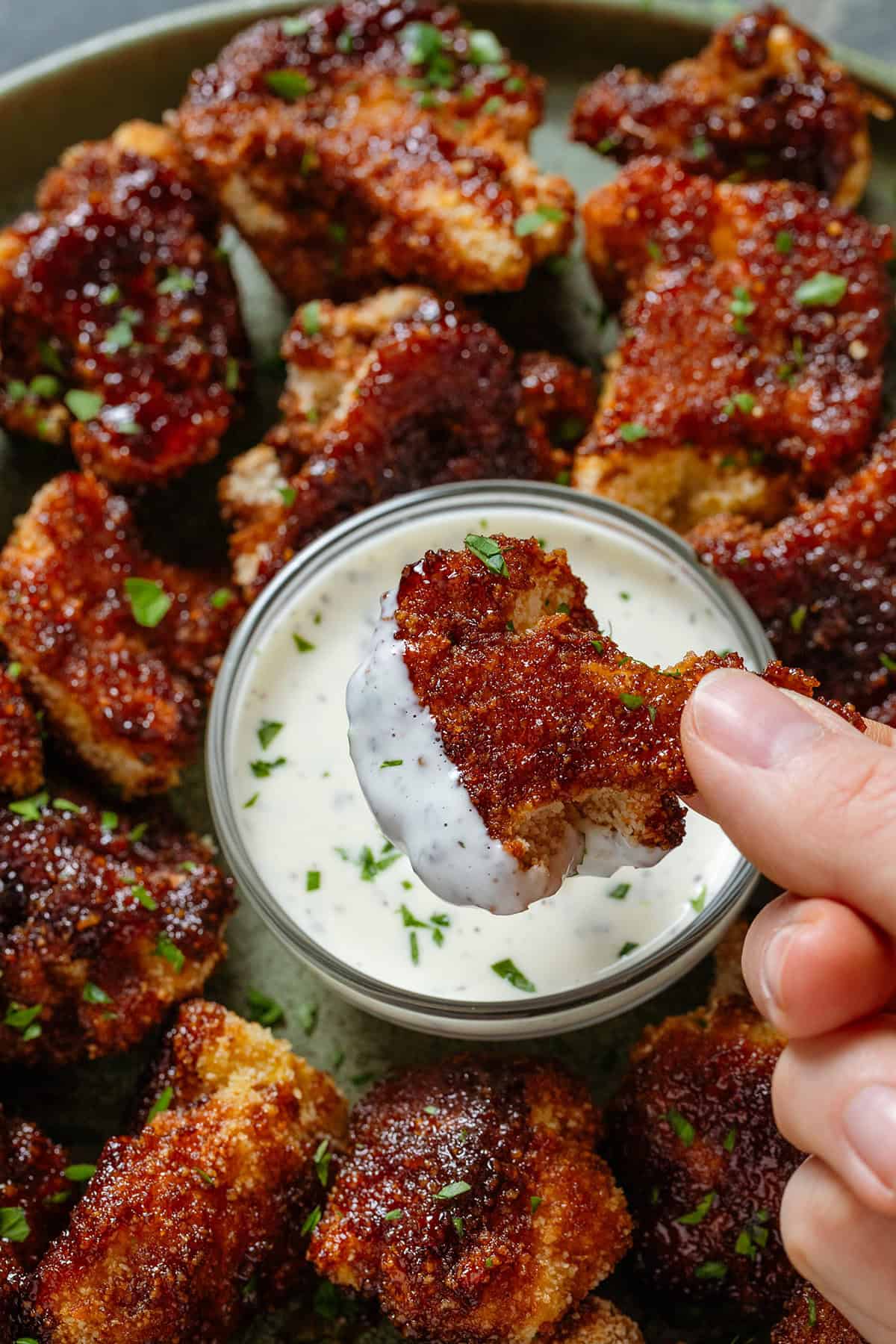 A hand dipping a crispy bbq cauliflower bite into white mayo sauce over a bowl of more cauliflower bites.