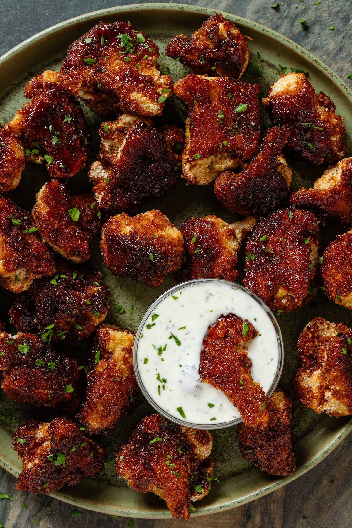 Baked breadcrumbed bbq cauliflower florets on a green plate with a small glass bowl of white dip in the middle with one floret dipped in it.
