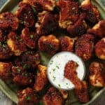 Baked breadcrumbed bbq cauliflower florets on a green plate with a small glass bowl of white dip in the middle with one floret dipped in it.